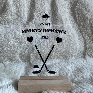 Könnte beinhalten: Klarer Acrylschild mit dem Text "IN MY SPORTS ROMANCE ERA" und Hockey-Grafiken. Das Schild wird von einem hellen Holzsockel getragen, vor einem flauschigen weißen Hintergrund.