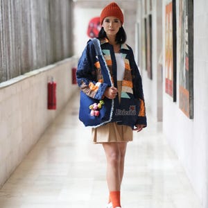 Puede incluir: Una mujer con una chaqueta vaquera con detalles de patchwork y un bolso tote a juego. La chaqueta tiene una parte superior blanca debajo, una falda color canela y un gorro naranja. El bolso tote tiene adornos florales y la palabra "Eriesta" es visible.