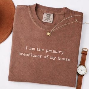 May include: A brown Comfort Colors t-shirt with the text "I am the primary breadloser of my house." A gold star pendant necklace and a brown leather watch are next to the shirt. A brown hat is partially visible in the upper left corner.