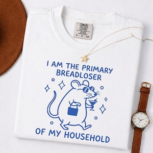 May include: White t-shirt with the text "I AM THE PRIMARY BREADLOSER OF MY HOUSEHOLD" in blue. A cartoon mouse wearing sunglasses and holding a purse and cocktail is printed below the text. A gold necklace and watch are also visible.