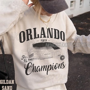 May include: Cream-colored sweatshirt with the word "ORLANDO" in large black letters above "-- 1989 --" and "Champions" in a script font. A detailed illustration of a stadium is featured below the text. The sweatshirt is a Gildan Sand.