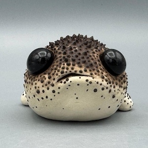 May include: A ceramic pufferfish sculpture with a cream-coloured body and a spiky, brown top. The sculpture has large, glossy black eyes and small, dotted details. The pufferfish is a whimsical decorative item.