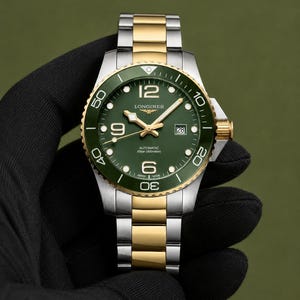 May include: A two-tone stainless steel and gold-tone Longines automatic watch with a green dial and green rotating bezel. The watch features a date window at the three o'clock position, luminous hands, and a metal link bracelet with gold-tone center links.
