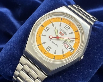 Peut inclure: Une montre-bracelet automatique Seiko vintage avec un boîtier et un bracelet argentés. Le cadran blanc est orné de détails orange, de chiffres noirs et d'un affichage jour-date. La montre est marquée "Japan Made".