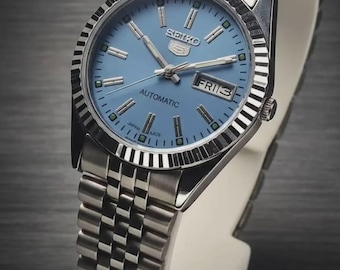 Peut inclure: Une montre-bracelet automatique Seiko 5 avec un cadran bleu clair, des index bâtons et des aiguilles argentés. Elle présente une lunette cannelée en acier inoxydable et un bracelet en métal à maillons. Un guichet jour-date est à trois heures.