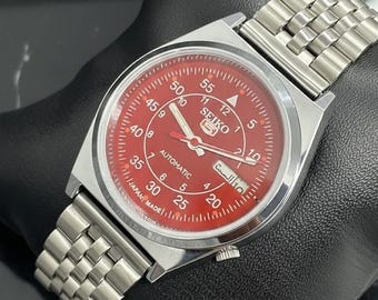 Peut inclure: Une montre-bracelet automatique Seiko 5 en acier inoxydable avec un cadran rouge vif. La montre est dotée d'un bracelet argenté, d'un affichage de la date et d'aiguilles lumineuses. Le cadran porte l'inscription "SEIKO 5 AUTOMATIC JAPAN MADE".