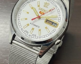 Peut inclure: Une montre automatique Seiko 5 argentée avec un cadran blanc. La montre présente des aiguilles et des index dorés, une trotteuse rouge et un affichage jour-date. Elle est dotée d'un bracelet en métal maillé.