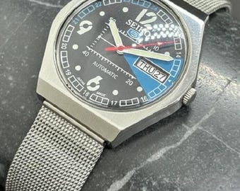 Peut inclure: Une montre-bracelet automatique Seiko argentée avec un cadran noir, des aiguilles blanches et rouges, et un bracelet en maille. Le cadran affiche le jour et la date, avec le texte "AUTOMATIC" et "SEIKO". La montre a un accent bleu et blanc sur le cadran.