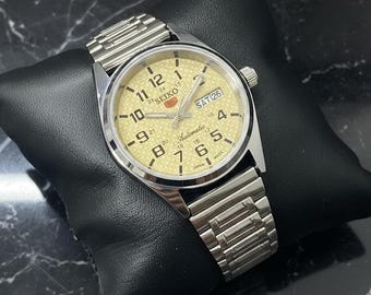 Peut inclure: Une montre automatique Seiko argentée avec un cadran doré clair. Le cadran affiche le jour et la date, avec des chiffres et des aiguilles noirs. La montre est sur un bracelet en métal argenté et repose sur un coussin noir.