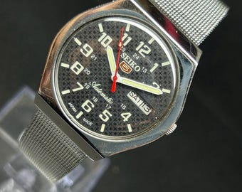 Peut inclure: Une montre-bracelet automatique Seiko 5 argentée avec un cadran noir texturé présentant un motif de croix répétitif. La montre possède un bracelet en maille d'acier inoxydable, des chiffres arabes blancs, une trotteuse rouge et un guichet jour-date.