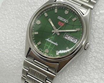 Peut inclure: Une montre-bracelet automatique Seiko argentée avec un cadran vert. Le cadran affiche le jour et la date, avec le jour indiquant "MER 6". La montre a un bracelet argenté et est posée sur un fond blanc.