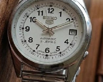 Peut inclure: Montre automatique Seiko argentée avec un cadran blanc. La montre présente des chiffres noirs, des aiguilles et le texte "AUTOMATIC 21 JEWELS". Le bracelet de la montre est argenté.