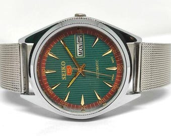 Peut inclure: Une montre-bracelet automatique Seiko 5 avec un cadran vert à lignes horizontales, des aiguilles et index dorés, et un guichet jour-date à trois heures. La montre possède un boîtier en acier inoxydable argenté et un bracelet en maille métallique.