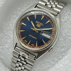 May include: A silver Seiko 5 automatic wristwatch with a blue dial and gold hands and markers. The watch face shows the day and date. The watch has a stainless steel strap.