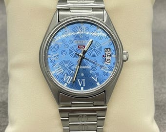 Peut inclure: Une montre automatique Seiko 5 en acier inoxydable avec un cadran bleu clair orné d'un motif en spirales et cercles. La montre a un bracelet argenté et un affichage de la date à 3 heures. La montre est présentée sur un coussin crème.