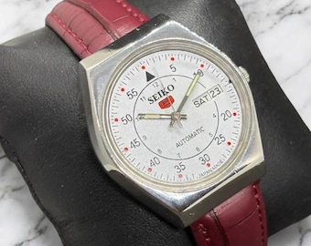 Peut inclure: Une montre automatique Seiko vintage avec un boîtier octogonal argenté et un cadran blanc. La montre est dotée d'un bracelet en cuir rouge et affiche le jour et la date. Le cadran comprend le nom de la marque et le texte "AUTOMATIC".