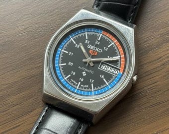 Peut inclure: Une montre-bracelet automatique Seiko vintage avec un cadran noir, des accents bleus et orange. La montre a un boîtier en acier inoxydable et un bracelet en cuir noir. Le jour est affiché comme "THU 20".