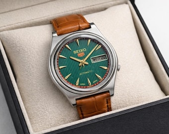 Peut inclure: Une montre-bracelet automatique Seiko avec un cadran vert à rayures verticales, des aiguilles et index dorés, et un guichet jour-date. La montre possède un boîtier en acier inoxydable argenté et un bracelet en cuir marron texturé.