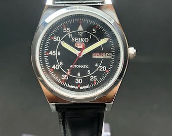 Peut inclure: Une montre-bracelet automatique Seiko 5 avec un cadran noir, des aiguilles argentées et un bracelet en cuir noir. Le cadran présente des chiffres arabes blancs pour les minutes et les heures, un guichet jour-date et l'inscription Japan Made.