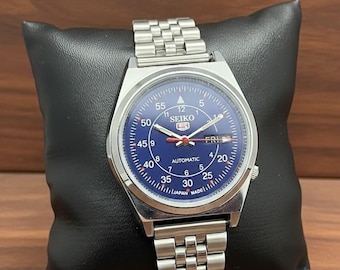 Peut inclure: Une montre automatique Seiko 5 en acier inoxydable avec un cadran bleu. La montre est dotée d'un bracelet argenté et est présentée sur un coussin noir. Le cadran comporte des chiffres, des aiguilles et le jour de la semaine blancs.