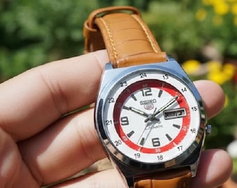 Peut inclure: Une montre automatique Seiko argentée avec un cadran blanc, des accents rouges et un bracelet en cuir marron. La montre affiche le jour et la date, avec le logo Seiko en haut. L'arrière-plan est flou, vert et jaune.