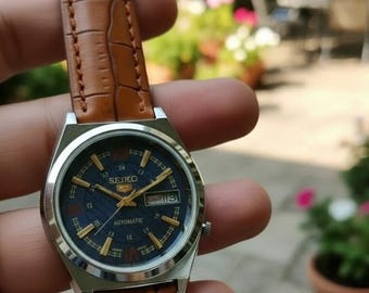 Peut inclure: Montre automatique Seiko 5 vintage avec cadran bleu et aiguilles et index dorés. Le boîtier est en acier inoxydable et le bracelet en cuir marron. Le cadran affiche le jour et la date.