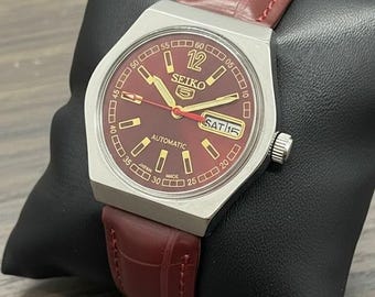 Peut inclure: Une montre automatique Seiko 5 avec un cadran bordeaux et un boîtier en acier inoxydable. La montre affiche le jour et la date et possède un bracelet en cuir bordeaux. La montre est présentée sur un coussin noir.