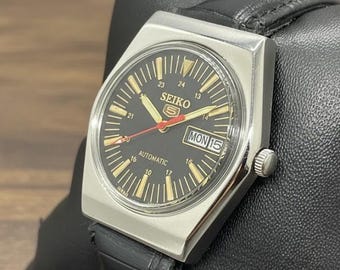 Peut inclure: Une montre automatique Seiko 5 vintage avec un cadran noir et des marqueurs dorés. La montre a un boîtier rectangulaire argenté et un bracelet en cuir noir. Le jour et la date sont affichés à 3 heures.