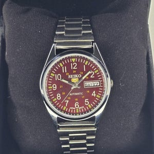 Puede incluir: Un reloj de pulsera automático Seiko 5 con correa de eslabones de acero inoxidable plateado y caja redonda. El reloj presenta una esfera de color rojo intenso con números arábigos blancos, ventana de día y fecha y agujas luminiscentes.