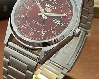 Peut inclure: Une montre-bracelet automatique Seiko en acier inoxydable avec un cadran bordeaux. La montre a un bracelet argenté et un cadran rond avec des détails blancs et dorés. Le cadran affiche le jour et la date, avec le mot "SEIKO" en haut.