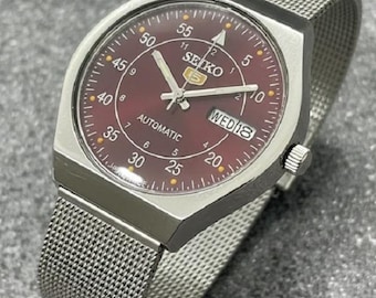 Peut inclure: Une montre automatique Seiko 5 en acier inoxydable avec un cadran bordeaux et un bracelet en maille. Le cadran affiche le jour et la date, avec des aiguilles et des marqueurs lumineux. La montre a un boîtier argenté et un bracelet en maille texturée.