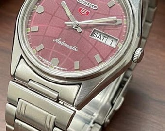 Peut inclure: Montre automatique Seiko 5 en acier inoxydable avec un cadran rouge à motif quadrillé. La montre affiche le jour et la date, avec "SAT 1" visible. Le bracelet est argenté.