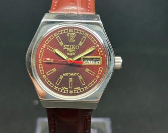 Peut inclure: Une montre-bracelet automatique Seiko 5 avec un cadran rouge profond, des index dorés et une trotteuse rouge. La montre présente un guichet jour-date à trois heures, un boîtier en acier inoxydable argenté et un bracelet en cuir rouge foncé.