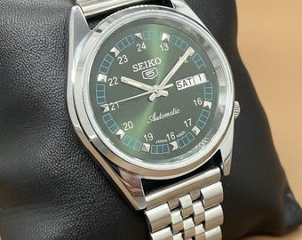 Peut inclure: Une montre automatique Seiko 5 argentée avec un cadran vert. La montre est dotée d'un bracelet en acier inoxydable et d'un affichage jour-date. Le cadran comprend le texte "SEIKO 5 Automatic" et le jour de la semaine.