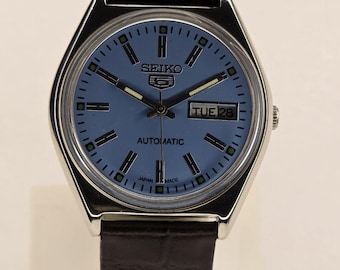Peut inclure: Une montre-bracelet automatique Seiko 5 avec un cadran bleu clair, des index argentés et un guichet jour-date à trois heures. La montre présente un boîtier argenté poli et un bracelet en cuir noir texturé avec une boucle.