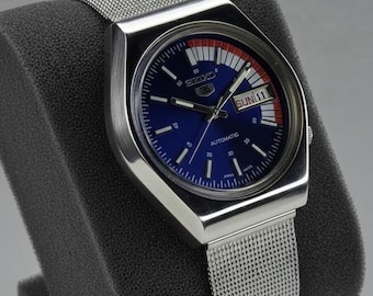 Peut inclure: Une montre automatique Seiko vintage avec un boîtier en acier inoxydable et un bracelet en maille. Le cadran est bleu avec des accents blancs et rouges, et affiche le jour et la date. La montre est présentée sur un support gris foncé.