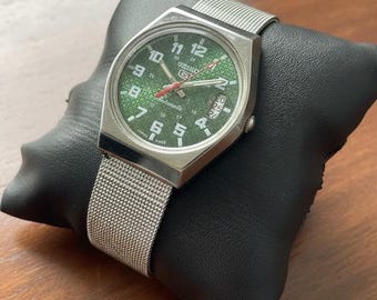 Peut inclure: Une montre Seiko argentée avec un cadran vert et un bracelet en maille. Le cadran de la montre présente des chiffres et des aiguilles blancs, avec un affichage de la date à 3 heures. La montre est présentée sur un coussin noir.