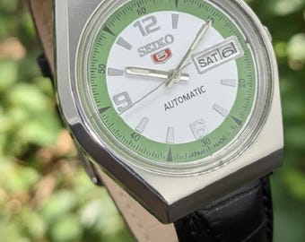 Peut inclure: Une montre automatique Seiko 5 vintage avec un boîtier argenté et un bracelet en cuir noir. Le cadran est blanc avec des accents verts, y compris les marqueurs de minutes et l'affichage jour/date. La montre affiche le jour "SAM 6".