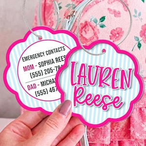 May include: Two scalloped, flower-shaped luggage tags with light blue and white vertical stripes and bright pink borders. One tag displays the name Lauren Reese in pink script, while the other lists emergency contacts for Mom and Dad.