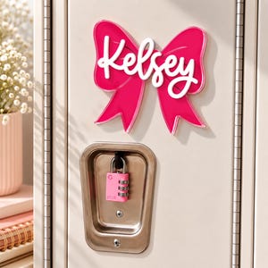 May include: A bright pink bow-shaped sign with the name Kelsey written in white script lettering, mounted on a light-colored locker door. Below the bow, a small pink combination padlock is secured within the recessed metal locker handle.