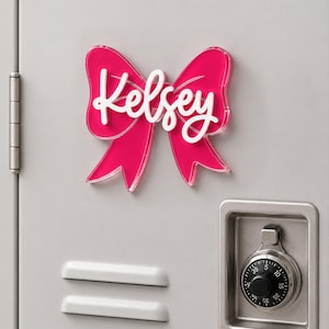 May include: A bright pink acrylic bow-shaped sign featuring the name Kelsey in white script lettering. The decorative piece is mounted on a light gray metal surface, positioned above a silver combination lock and two horizontal ventilation slats.