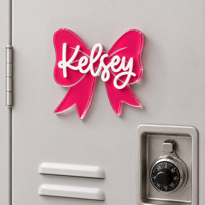 May include: A bright pink acrylic bow-shaped sign featuring the name Kelsey in white script lettering. The decorative piece is mounted on a light gray metal surface, positioned above a silver combination lock and two horizontal ventilation slats.