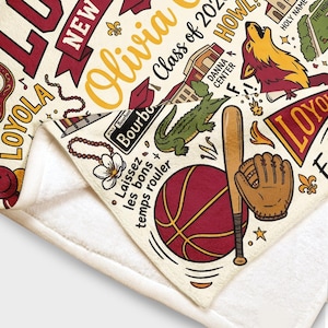 Puede incluir: Manta color crema con un dise&ntilde;o burdeos y dorado. Presenta el nombre "Olivia", "Class of 2023" e ilustraciones que incluyen un lobo, un caim&aacute;n, b&eacute;isbol y baloncesto. Incluye el texto "Laissez les bons temps rouler".