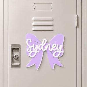 May include: A light purple bow-shaped sign featuring the name Sydney in white script lettering. The decorative piece is attached to a light gray metal locker door with a silver handle and a rectangular ventilation slot above the bow.