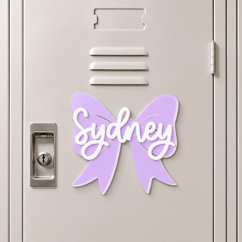 May include: A light purple bow-shaped sign featuring the name Sydney in white script lettering. The decorative piece is attached to a light gray metal locker door with a silver handle and a rectangular ventilation slot above the bow.