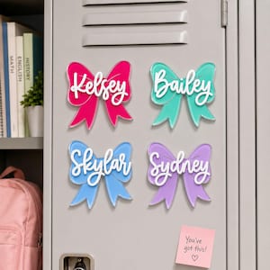 May include: Four bow-shaped magnets in pink, teal, light blue, and purple, each featuring a white script name: Kelsey, Bailey, Skylar, and Sydney. A small pink square sticky note with the text You're got this! and a heart symbol is below.