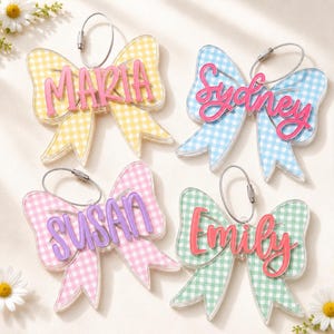 May include: Four bow-shaped acrylic bag tags with a gingham pattern. The yellow bow features the name MARIA in pink, the blue bow features SYDNEY in pink, the pink bow features SUSAN in purple, and the green bow features EMILY in red.