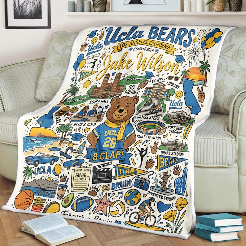 Puede incluir: Una manta blanca de UCLA Bears con un dise&ntilde;o colorido con el texto "UCLA BEARS" y "Jake Wilson". La manta incluye ilustraciones de lugares emblem&aacute;ticos, una mascota de oso y el texto "Class of 2026".