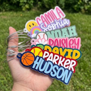 May include: A collection of personalized acrylic bag tags featuring various icons including a smiley face, rainbow, baseball, flower, turtle, softball, and basketball. Each tag displays a name in bold, colorful lettering attached to a metal cable.