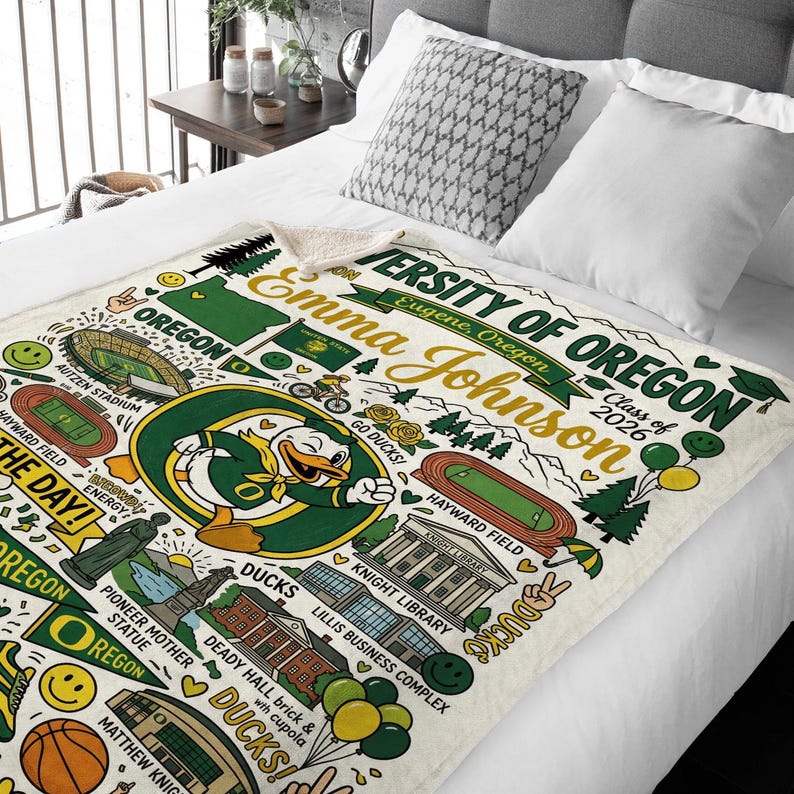 Puede incluir: Una manta crema de la Universidad de Oreg&oacute;n con un gran gr&aacute;fico de la mascota Oregon Duck, el texto "Emma Johnson" y "Class of 2026". La manta tambi&eacute;n incluye ilustraciones de los puntos de referencia del campus.